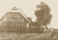 Stare sepiowe zdjęcie szkoły, budynek po lewej stronie zdjęcia, po prawej wysokie drzewo, przed zabudowaniami drewniany płot